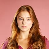 Une jeune femme aux longs cheveux roux et à l'expression sérieuse pose sur un fond rose, vêtue d'un haut marron.