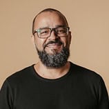 Un homme aux cheveux courts, à la barbe fournie et aux lunettes sourit, vêtu d'une chemise noire sur un fond beige uni.