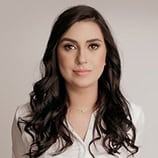 Une femme aux longs cheveux bruns ondulés et à l'expression neutre, vêtue d'une chemise blanche boutonnée, se tient sur un fond clair uni.