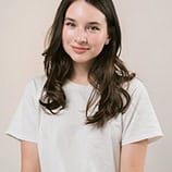Une jeune femme aux longs cheveux bruns, vêtue d'un simple tee-shirt blanc, se tient devant un fond clair et sourit légèrement.