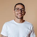 Homme aux cheveux courts, à la barbe taillée et aux lunettes, portant un t-shirt blanc, debout devant un fond beige uni.