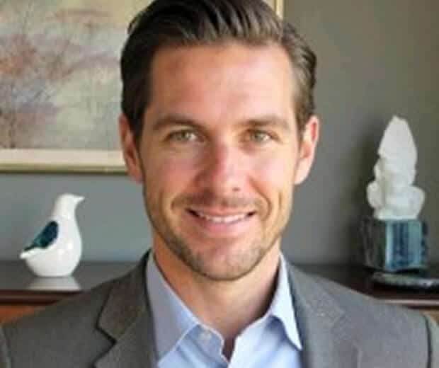 Un homme aux cheveux bruns courts et à la barbe taillée, vêtu d'une chemise bleu clair et d'un blazer gris, sourit devant une étagère contenant des objets décoratifs.