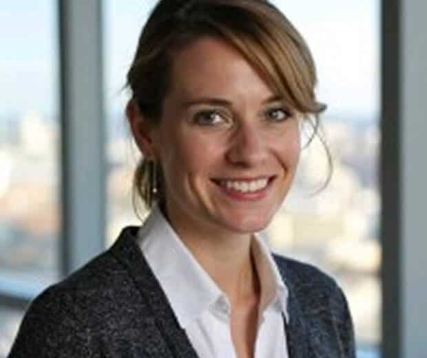 Une femme aux cheveux châtains clairs coiffés en queue de cheval, vêtue d'une chemise blanche et d'un pull sombre, sourit devant de grandes fenêtres de bureau offrant une vue sur la ville.