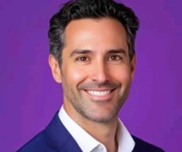 Homme souriant aux cheveux bruns portant un blazer bleu marine et une chemise blanche, posé devant un fond violet uni.